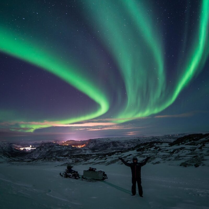 Northern Lights streaking across a dark night sky in swirls and cruves of green, with a person standing underneath next to snowmobiles