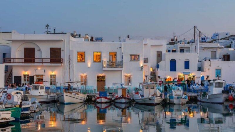 Illuminated white buildings and fishing boats reflected in a calm Greek harbor at dusk.