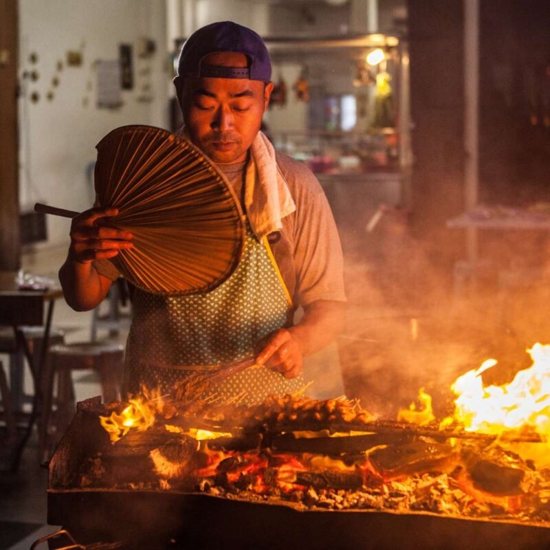 A man grilling food over a fire with a hand fan on luxury Southeast Asia holidays.