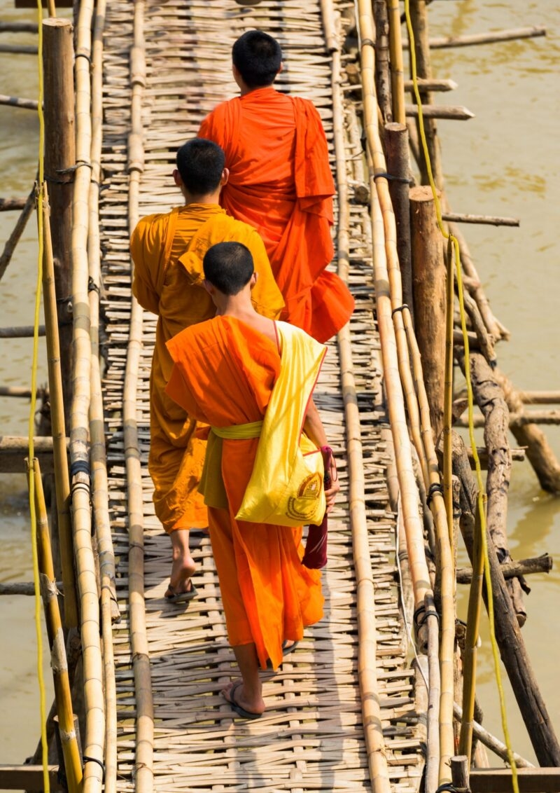 Three monks wearing orange robes cross a rustic bamboo bridge, a serene scene from luxury Laos holidays.