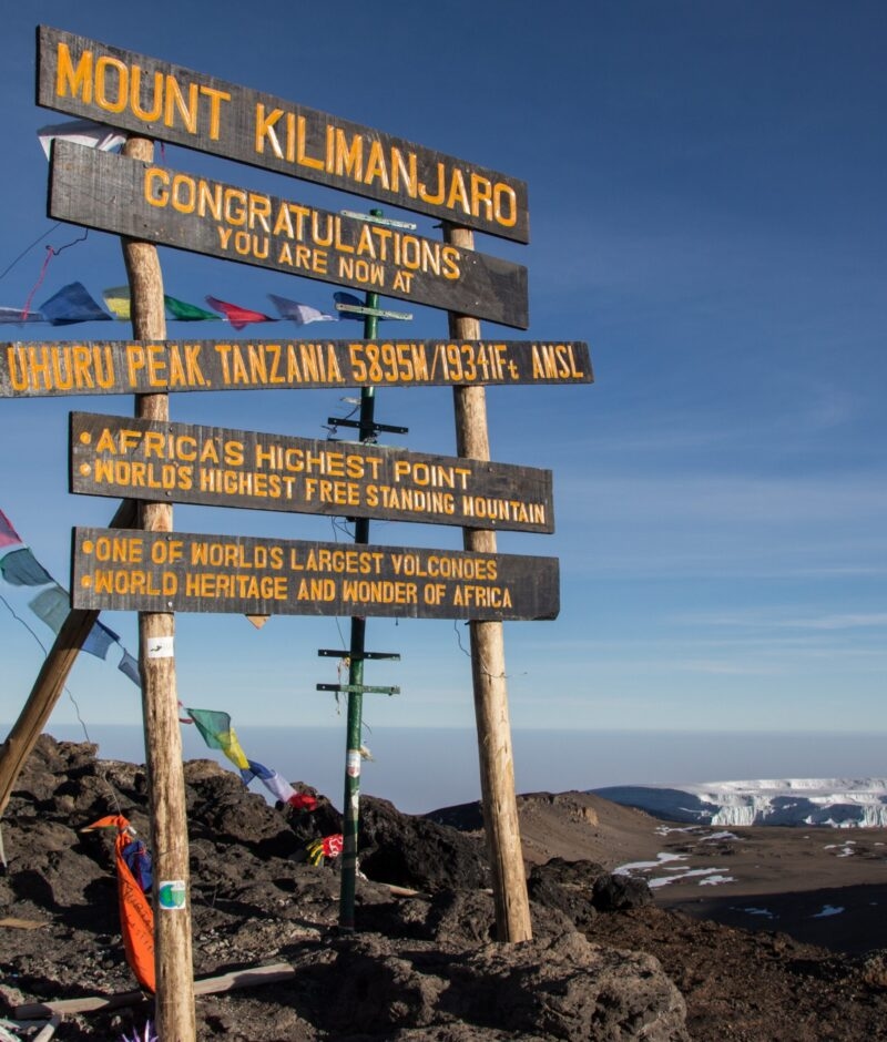 A wooden summit sign at Mount Kilimanjaro Uhuru Peak with prayer flags and a glacier in the distance.