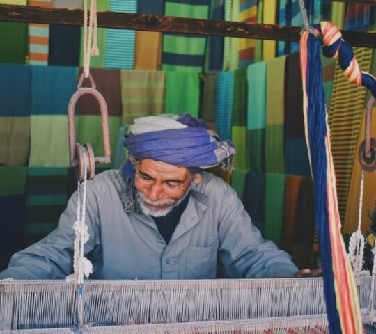 A man working at a traditional wooden loom with rows of colorful green and blue fabrics in the background.