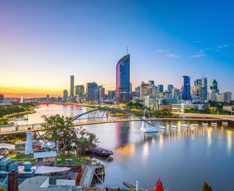 An evening view of a city skyline by a river with a lit-up arched bridge and a small lighthouse.
