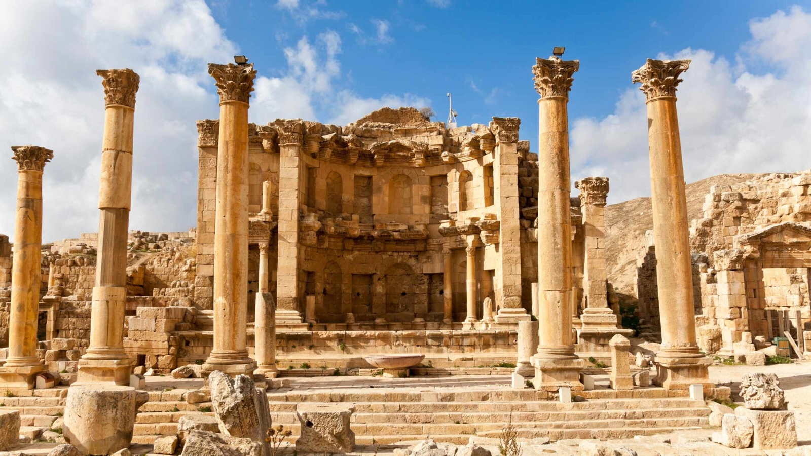 Ancient Roman stone ruins with tall Corinthian columns and a central fountain structure against a blue sky.