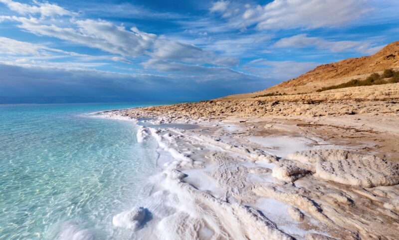 The Dead Sea shoreline featuring bright white salt formations, clear turquoise water, and rocky, beige hills under a blue sky.