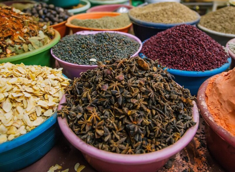 Close-up of various spices and grains, including star anise, lentils, dried garlic flakes, and red seeds, displayed in market bowls.
