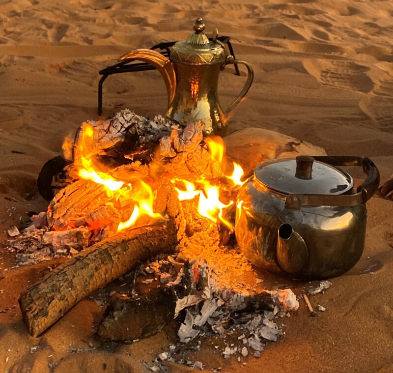 Two brass kettles, a traditional coffee pot and a round tea pot, sit beside a small campfire on the desert sand.