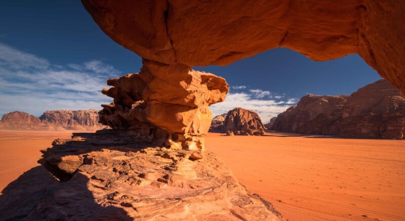 Natural sandstone rock arch formation frames the reddish-orange desert and mountains of Wadi Rum under a deep blue sky.