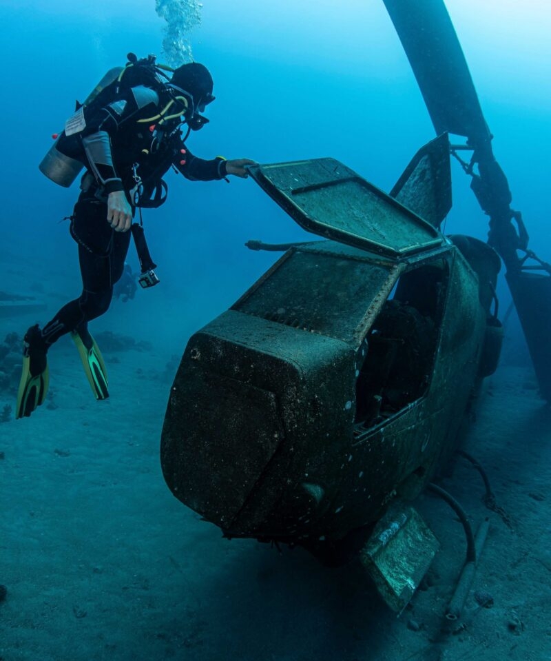 A scuba diver examines the cockpit of a dark, rusty, submerged military helicopter wreck on a sandy ocean floor.