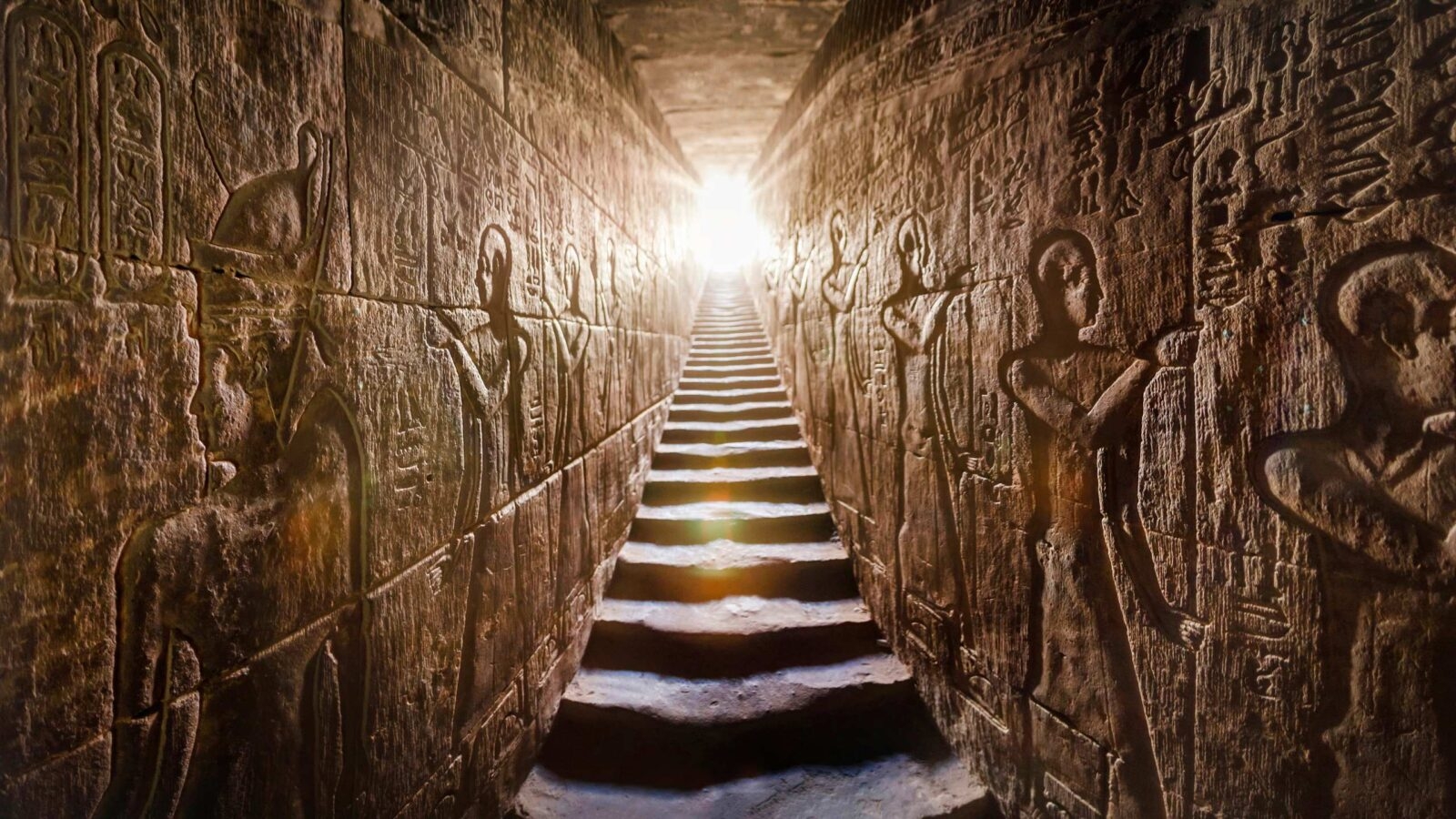 A stone stairway between two walls featuring intricate Egyptian reliefs leading up toward a bright light.