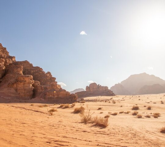 A wide, bright view of a red-sand desert with tall, rugged sandstone mountains under a clear blue sky. Luxury Middle East trips.