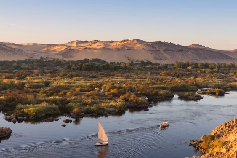 An aerial view of the wide Nile River flowing between green islands and sandy desert hills, with a sailboat, on luxury Middle East trips.