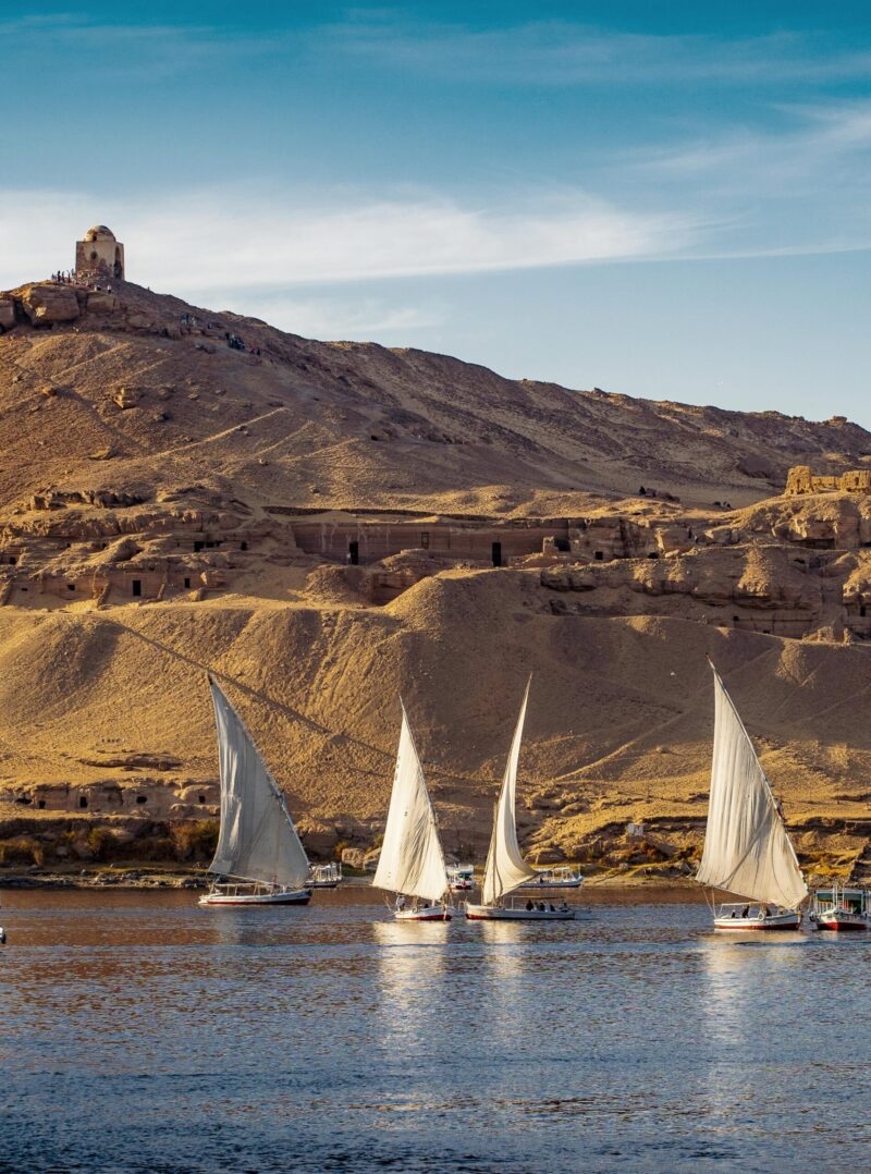 Several traditional white-sailed feluccas on the Nile River, passing a large, tomb-filled, sandy hill. A luxury Middle East tour scene.