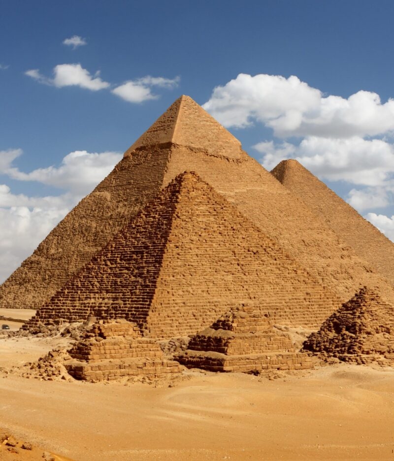 Three immense, ancient pyramids rise from the sandy desert ground under a blue, cloudy sky. An essential sight for a luxury Middle East vacation.