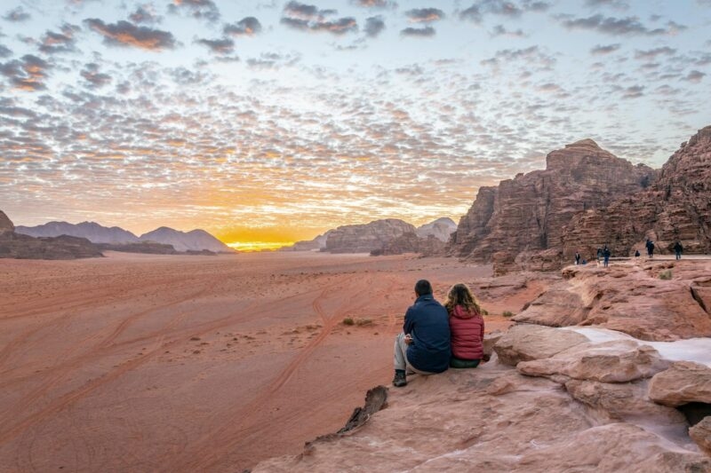 A man and woman sit on a rocky ridge, viewing a vast desert valley and mountains at sunset, perfect for luxury Middle East trips.