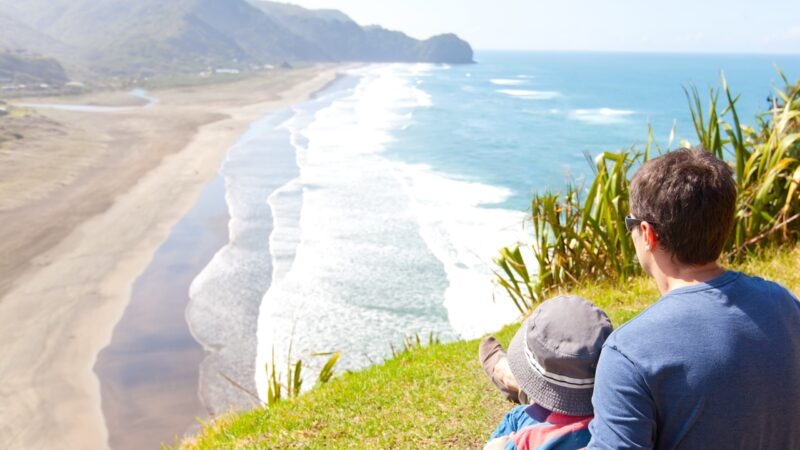back view of family sitting at the top of lion rock at piha beach, new zealand
