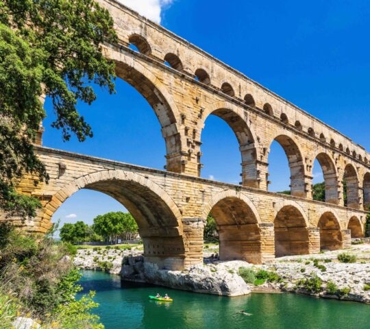 A high-angle view of the three-tiered Pont du Gard Roman aqueduct crossing a river with a green kayak below.