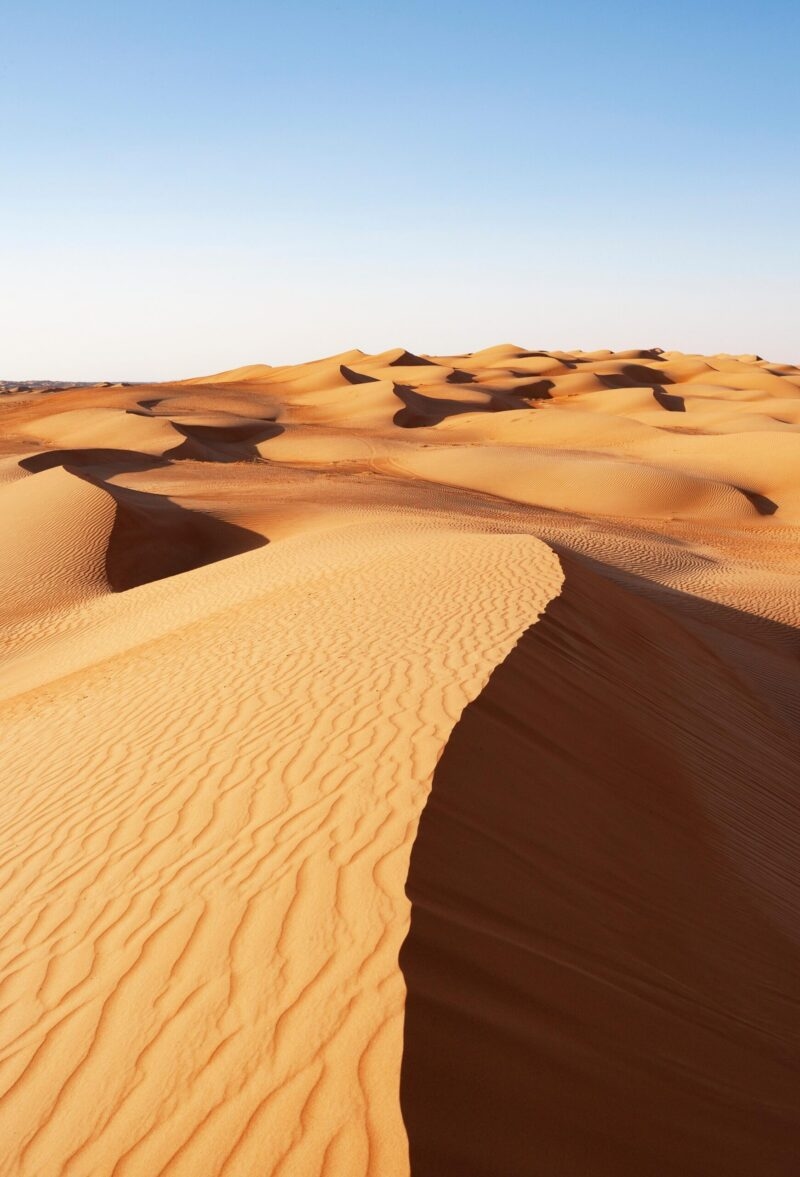 A vertical shot of towering, curving, golden sand dunes casting dark shadows beneath a clear blue sky. Luxury Middle East vacations.