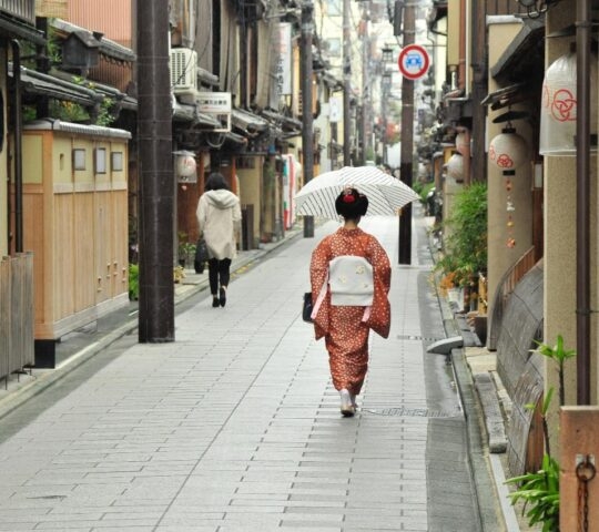 Back view of a person in a traditional kimono walking with an umbrella through a narrow paved alleyway in a city.