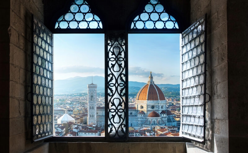 Looking through an open arched window at a city featuring a large dome and a tall square tower.