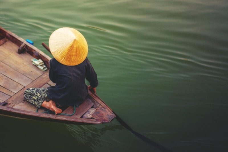 A person wearing a traditional conical hat rows a small wooden boat across dark, rippling water.