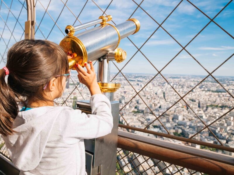 Little girl looking the cityscape of Paris from the top floor of the Eiffel Tower using the telescope