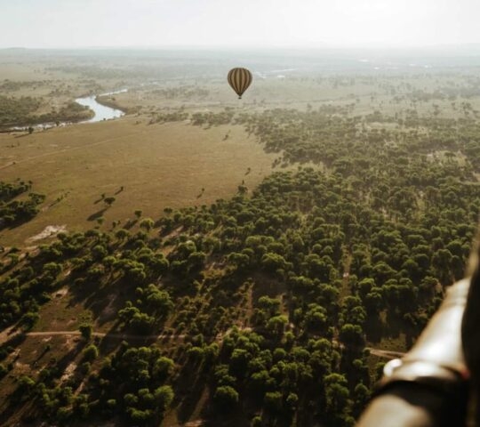 Aerial view of a hot air balloon flying over a wide grassy plain with a river and forest at sunrise.