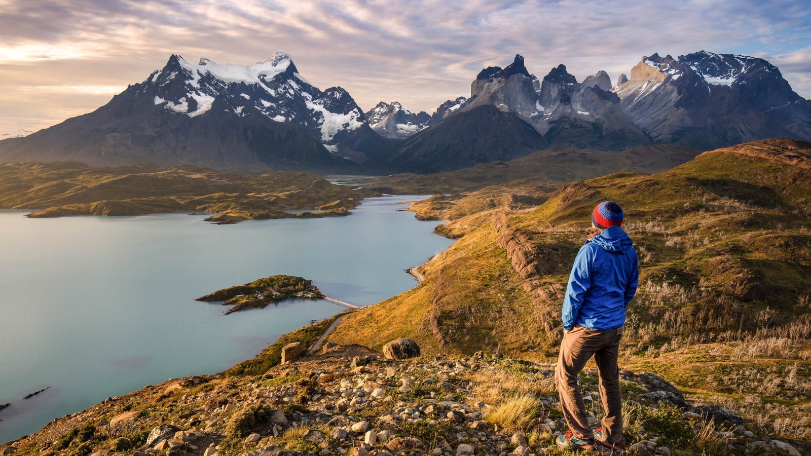Back view of a person in a blue jacket standing on a hill looking at a wide lake and sharp mountain peaks.