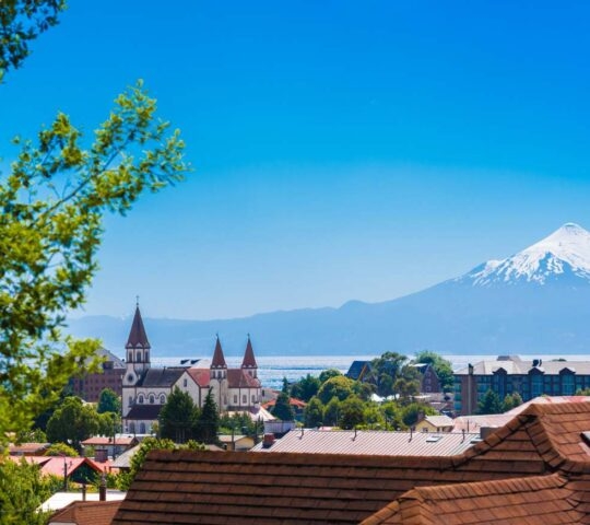 A white church with red steeples overlooking a town and water, with a perfectly conical snow-capped volcano behind.