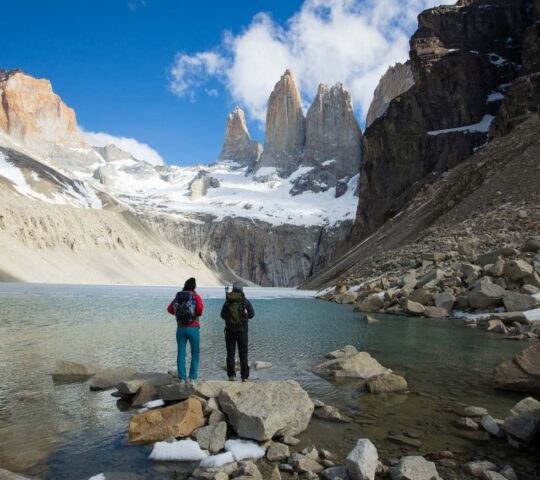 Two people with backpacks stand by a clear blue mountain lake facing jagged granite peaks and snow.