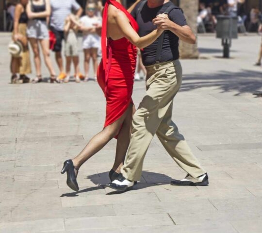 A woman in a red dress and a man in tan pants dancing tango in an open paved square.