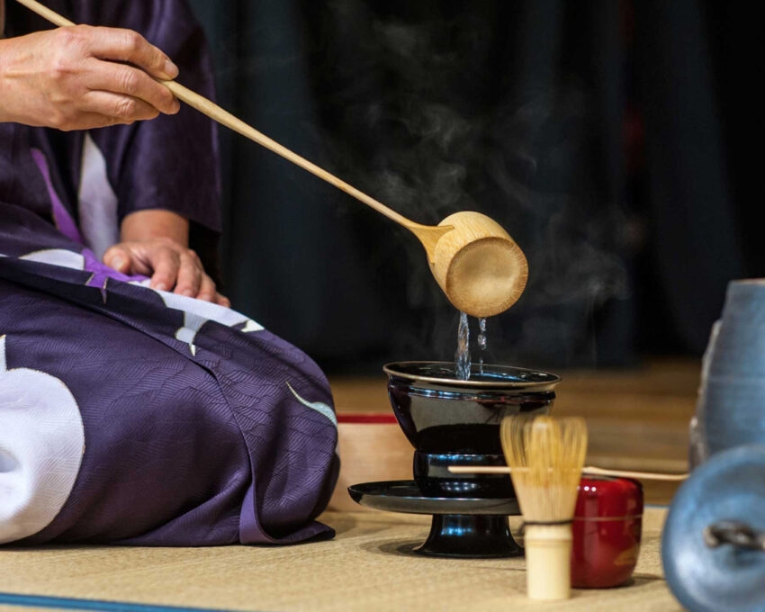 Close-up of a tea ceremony showing water poured from a wooden ladle into a black bowl on a tatami mat.