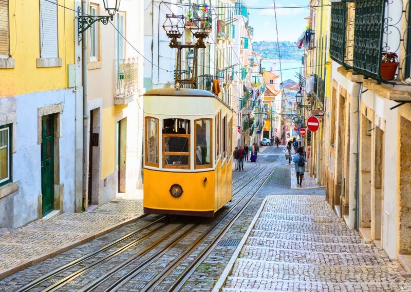 A yellow funicular tram travels down a steep street between tall, colorful buildings toward the sea in the distance.