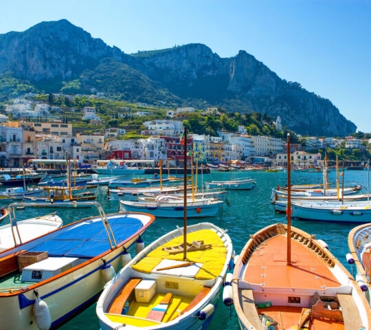 Several colorful wooden boats moored in a blue harbor with a large mountain and coastal town in the background.