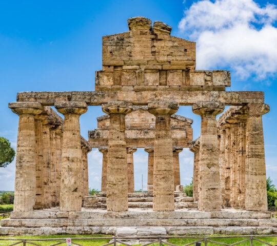 Large ancient stone temple with classic Doric columns standing against a bright blue sky with scattered clouds.