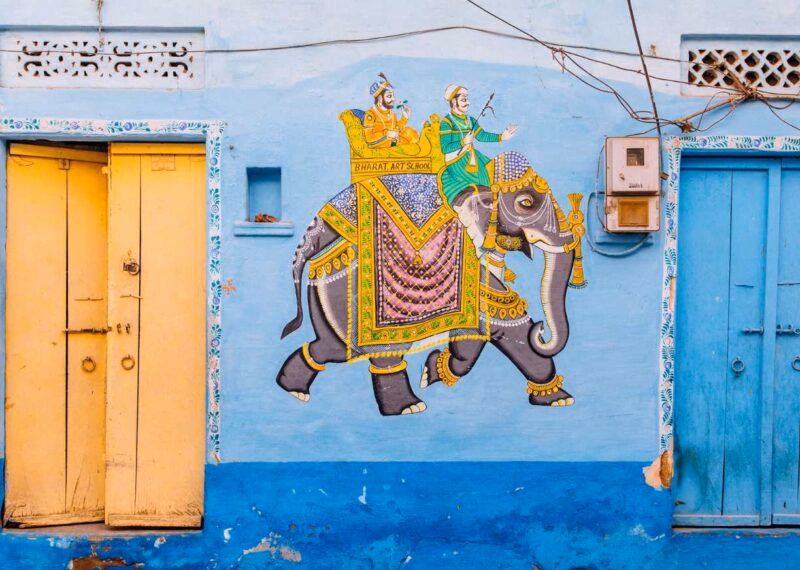 A painted mural on a blue wall, showing an elephant with people riding on top of it