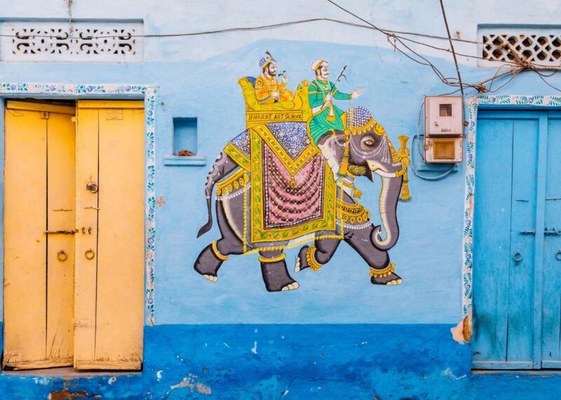 A painted mural on a blue wall, showing an elephant with people riding on top of it
