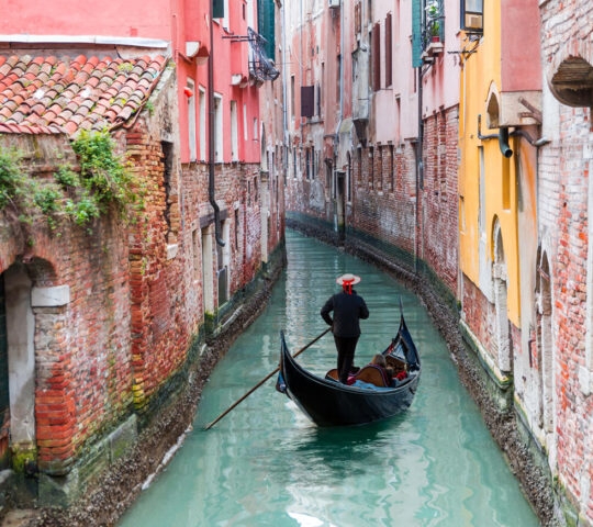 A man in a hat rows a black gondola down a narrow water canal between tall red and orange brick houses.