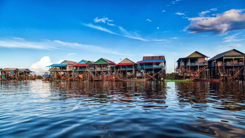 A row of colorful stilt houses in a floating village on a calm body of water reflecting the blue sky.