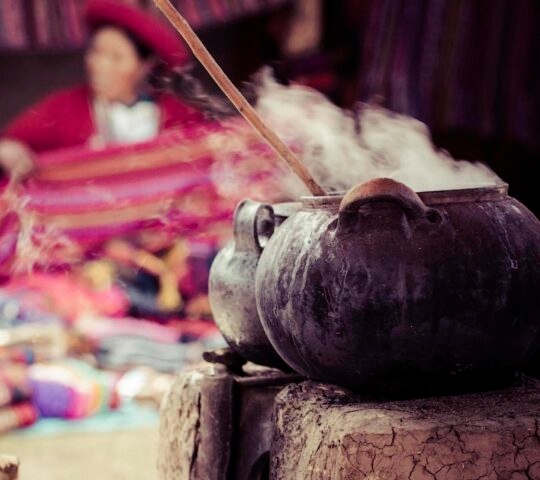 Steaming pot used in the traditional process of dyeing alpaca fur to make yarn for weaving with two Patacanchan people with their finished weaves out of focus in the background, Sacred Valley, Peru