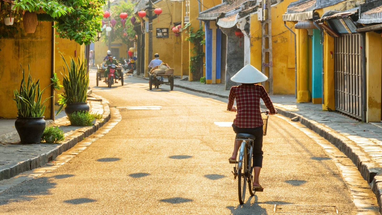 A cyclist wearing a conical hat rides down a sunlit street between yellow buildings in an old town.