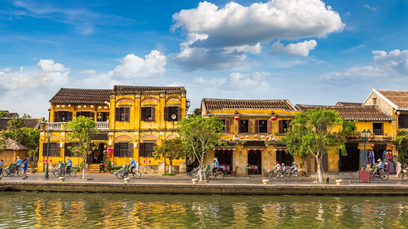A row of bright yellow historic buildings along a riverbank with reflections in the water under a blue sky.