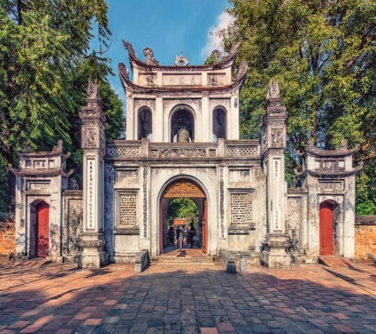 A large, ornate white stone gate with red doors and arched entryways, surrounded by tall green trees.