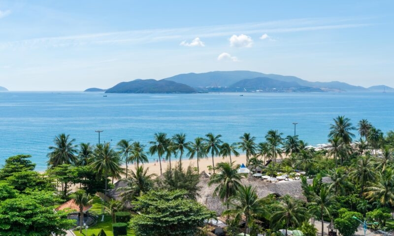 Scenic view of a sandy beach with palm trees and huts, looking out toward blue sea and mountains.