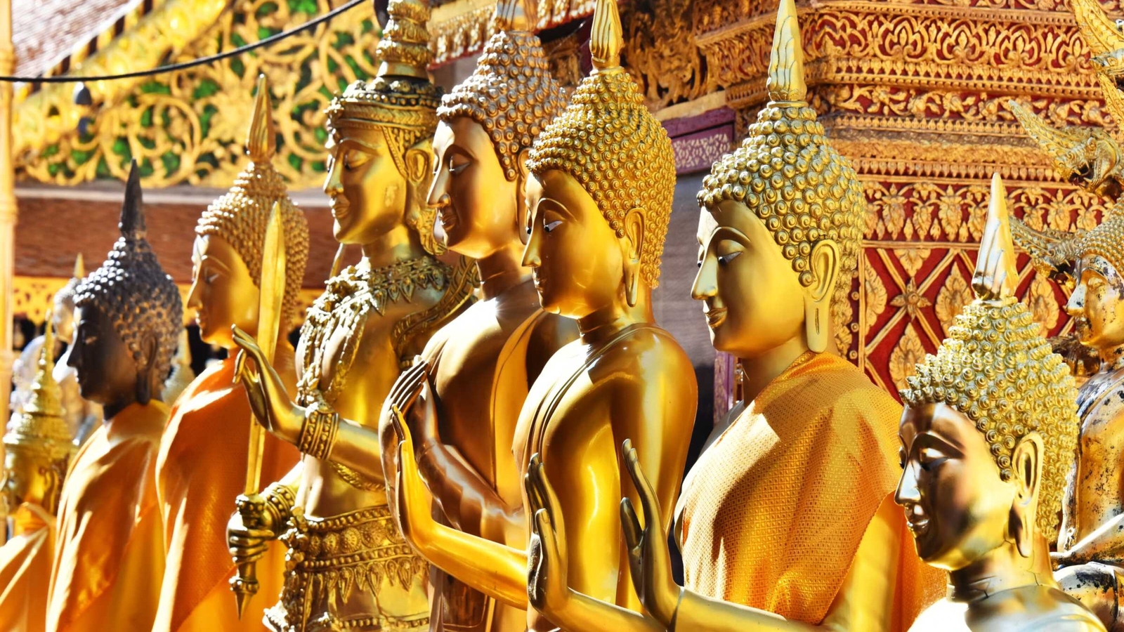 A close-up side view of several golden Buddha statues lined up in front of an intricately carved temple wall.