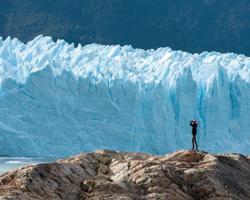 Small figure of a person standing on rocks in front of a giant, textured blue ice wall of a glacier.