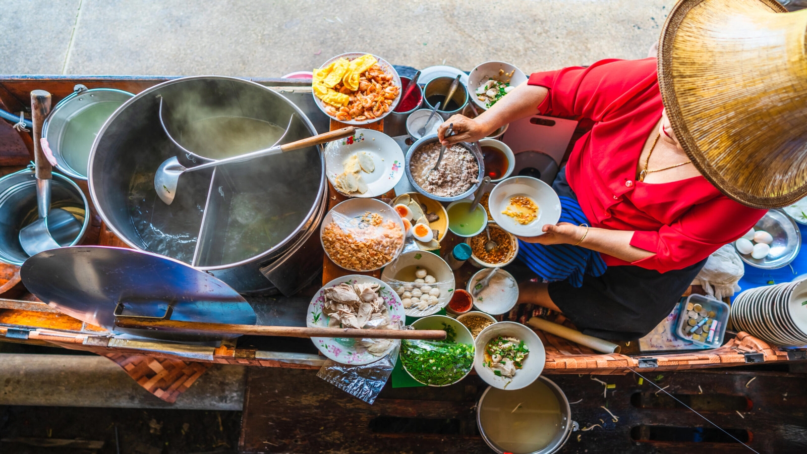 Overhead view of a food vendor in a boat surrounded by many bowls of ingredients and steaming pots.