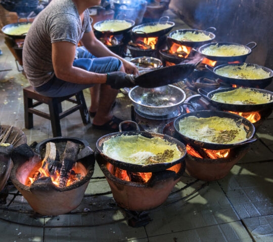 A person cooking several thin yellow pancakes on small round stoves with glowing orange embers.
