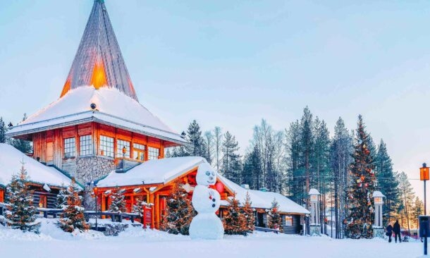 A giant snowman in front of a lit wooden cabin with a tall steeple in a snowy forest.