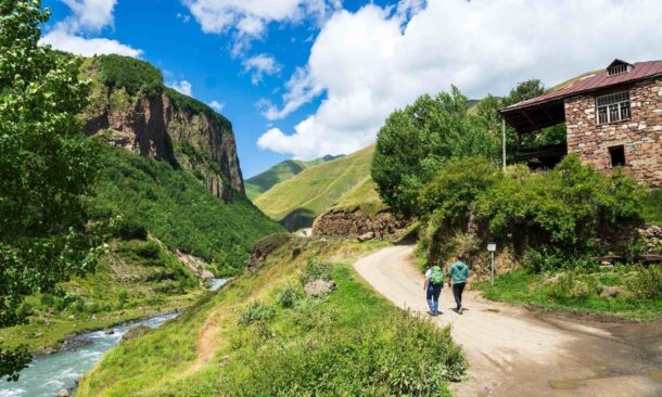 Two people hiking on a dirt road in a green mountain valley next to a stone house and a flowing river.
